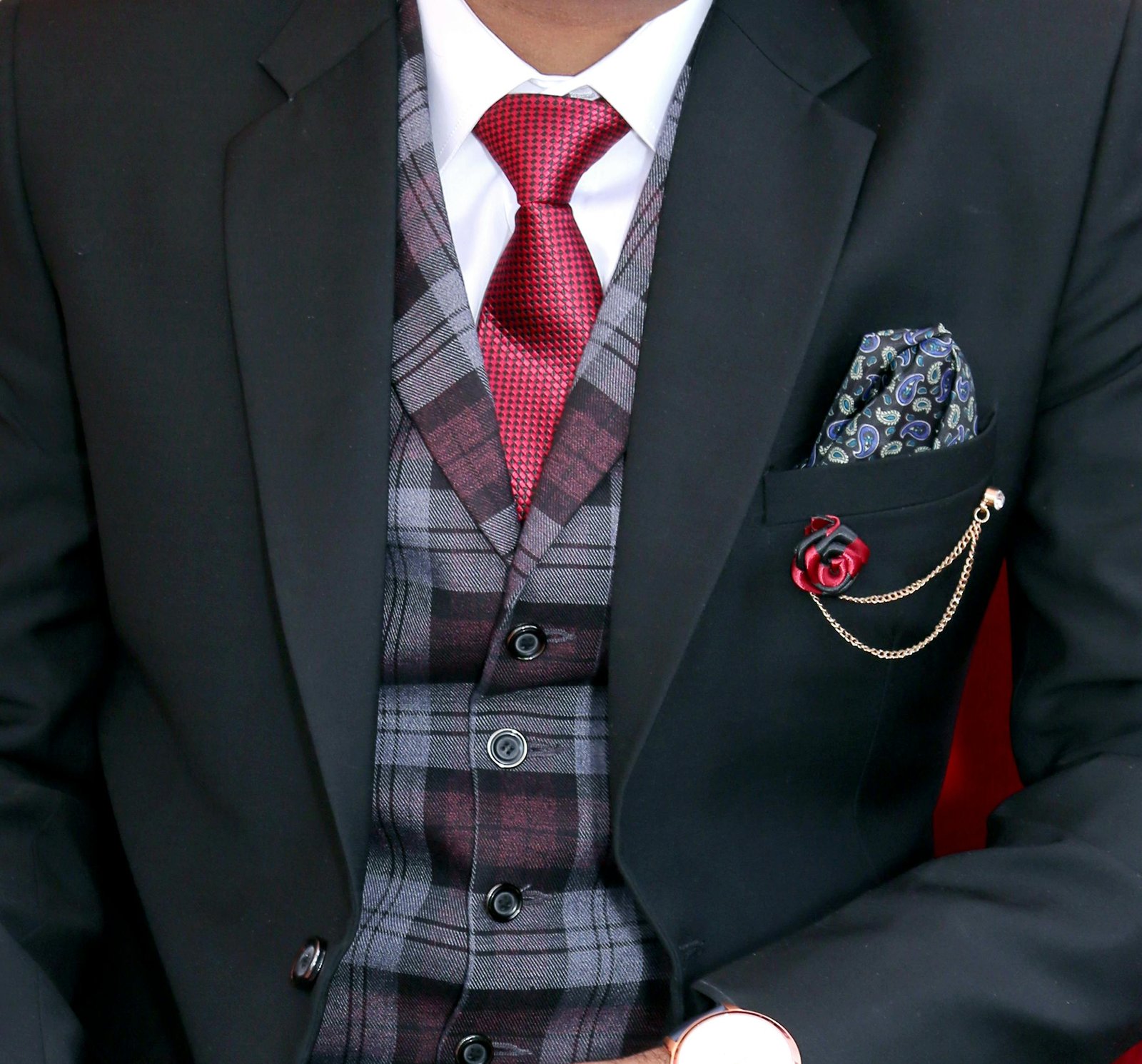 Close-up of a man in an elegant black suit with a red tie and plaid vest, showcasing formal menswear.