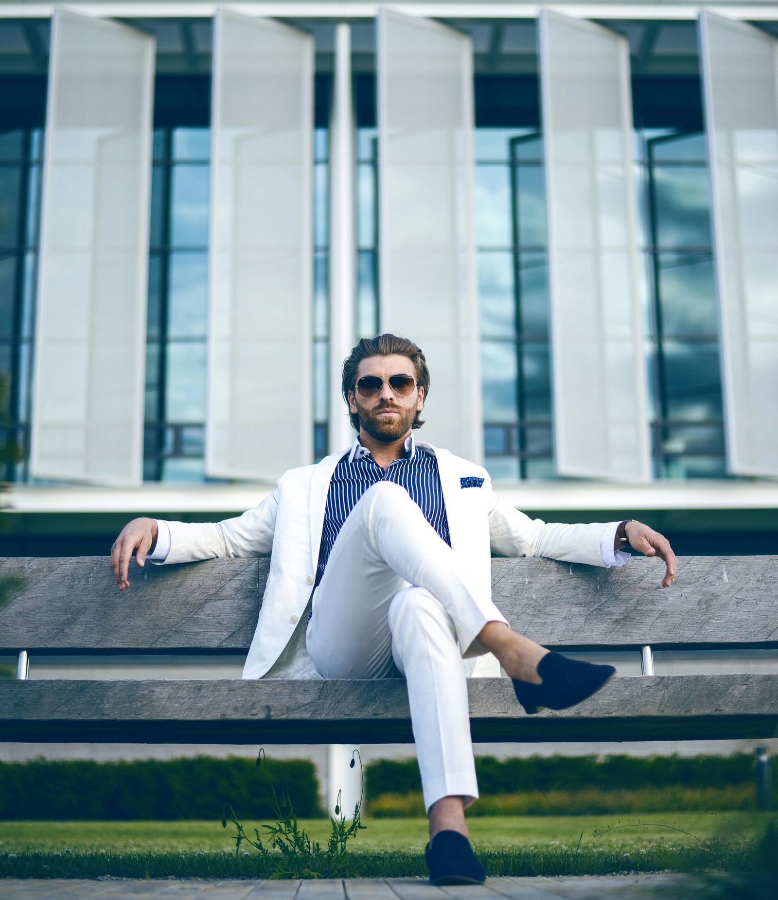 Fashionable man in white suit sitting confidently on a bench outdoors, embodying elegance and modern lifestyle.